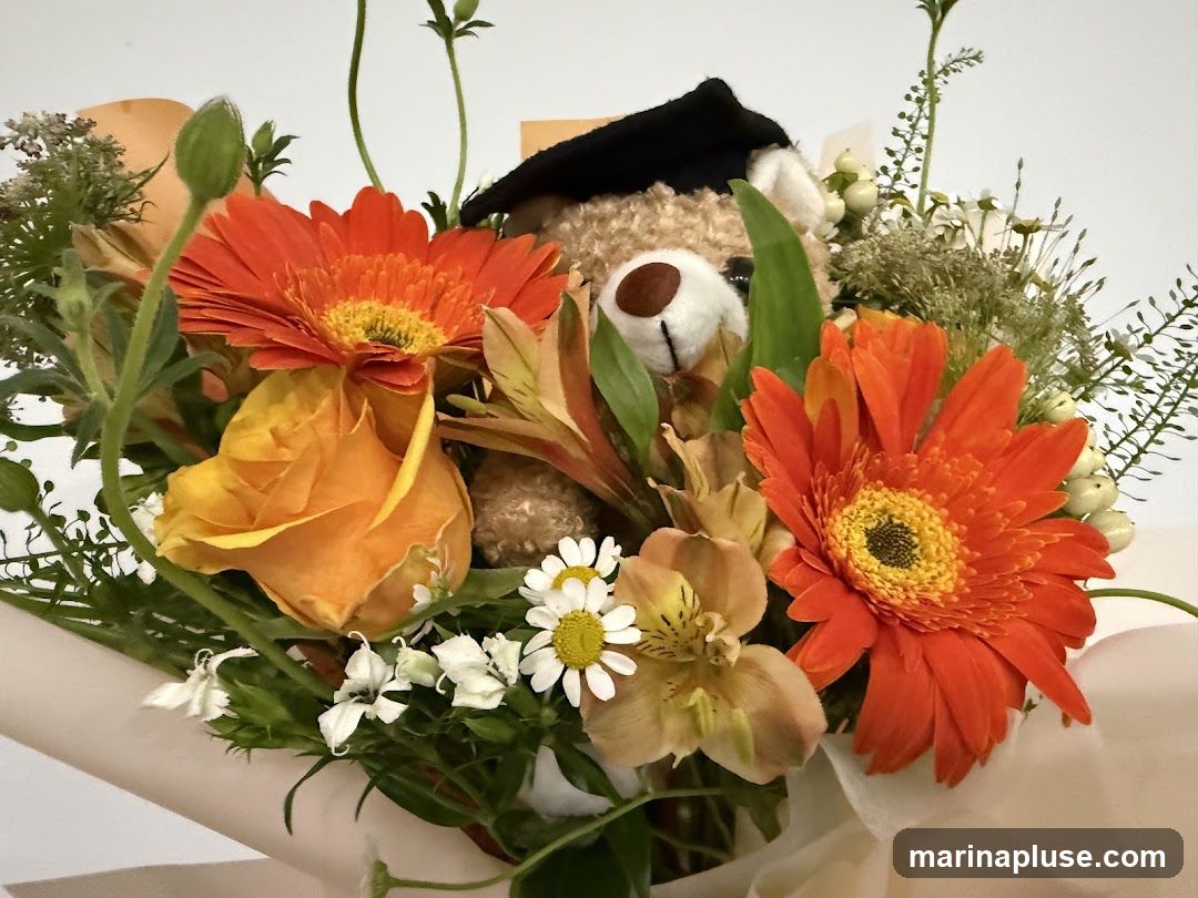 A close-up shot of a bouquet featuring a teddy bear wearing a graduation cap, surrounded by vibrant orange gerbera daisies, a yellow rose, and other assorted flowers and greenery. The bouquet is wrapped in light-colored paper.