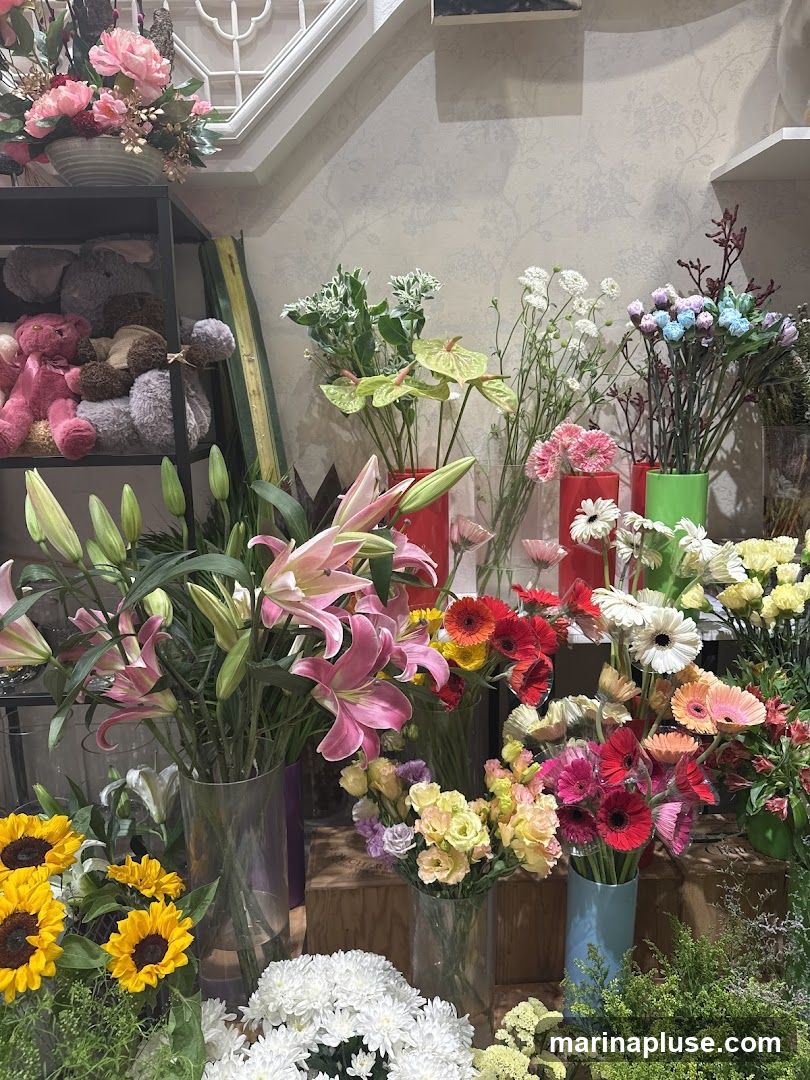 A vibrant display of various flowers, including lilies, sunflowers, gerbera daisies, and chrysanthemums, arranged in vases and on shelves. Stuffed animals are also visible on a shelf in the background.