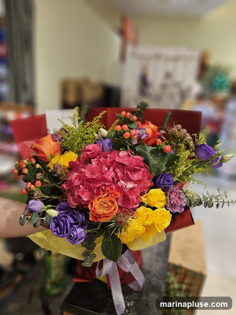A close-up shot of a colorful bouquet of flowers, featuring a large pink hydrangea as the centerpiece, surrounded by orange roses, yellow roses, purple lisianthus, and other assorted greenery and berries. The bouquet is wrapped in red and yellow paper and tied with a light purple ribbon. The background is blurred, showing hints of an indoor setting with shelves and possibly other floral arrangements.