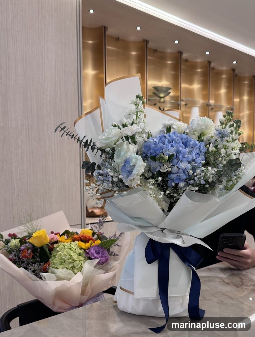 Two flower bouquets are displayed on a marble surface. The bouquet in the foreground is larger and features blue hydrangeas, white roses, and delicate white filler flowers, wrapped in white and light blue paper with a navy blue ribbon. The bouquet in the background is smaller and contains yellow, orange, and purple flowers, with a green hydrangea, wrapped in light pink paper. The background shows a modern interior with wood-paneled walls and gold-accented shelving with decorative items. A person's hand holding a smartphone is visible on the right side.