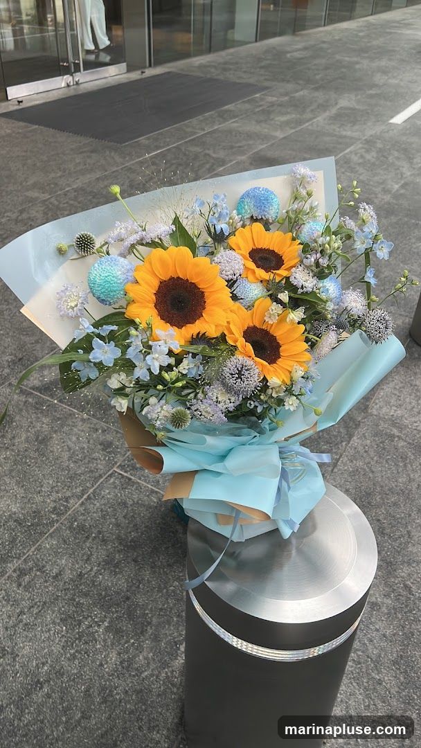 A bouquet of sunflowers, blue thistles, and small blue and white flowers is wrapped in light blue paper and tied with a ribbon. The bouquet is placed on a metal bollard on a stone-tiled sidewalk in front of a building with glass doors.