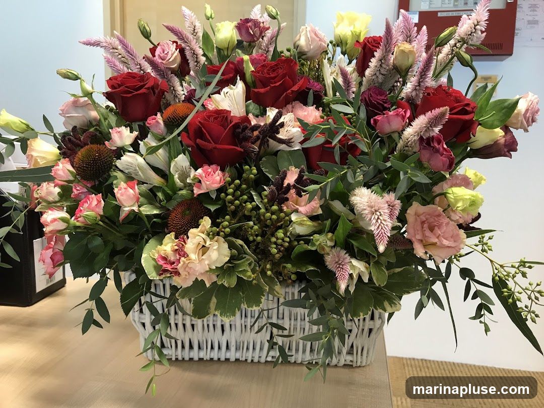 A large, lush flower arrangement in a white wicker basket sits on a wooden desk. The arrangement features a variety of flowers including red roses, pink roses, light pink lisianthus, white alstroemeria, and feathery pink celosia. Green foliage and eucalyptus branches add depth and texture. In the background, a wall with a fire alarm panel is visible.