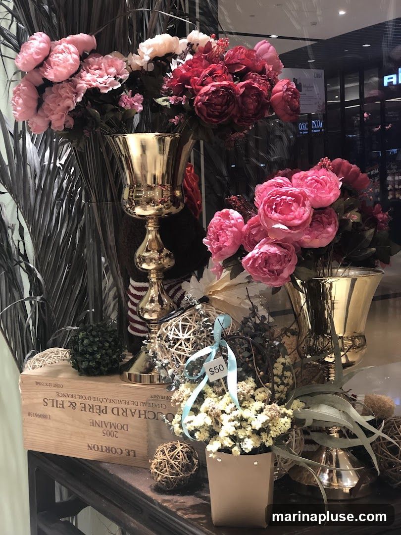 A display of artificial flowers in gold vases, with decorative orbs and a wooden crate. A price tag of $50 is visible on one of the arrangements.
