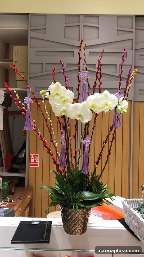 A decorative arrangement of white orchids and pussy willows with purple tassels sits in a textured gold pot on a counter. Behind the arrangement, a wall features a geometric pattern. To the left, a fire extinguisher sign is visible on a wooden panel. A clipboard and pen are on the counter in front of the arrangement.