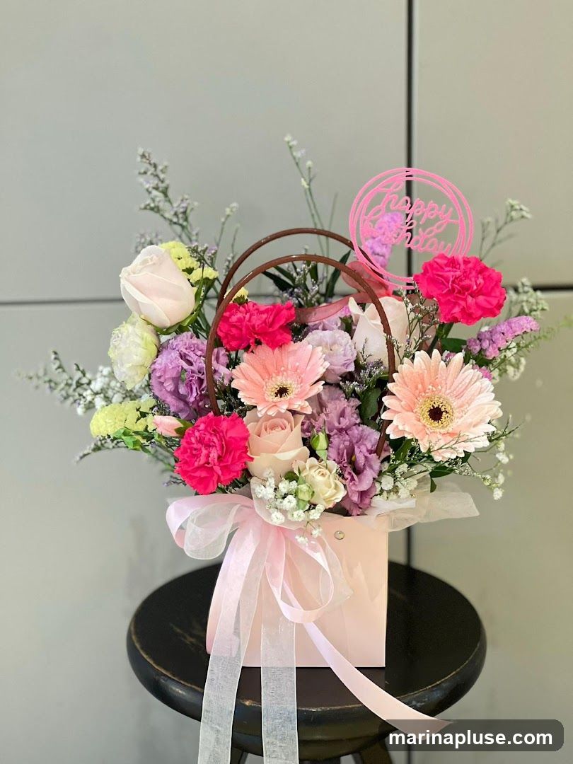 A pastel pink flower arrangement in a matching pink box, adorned with a sheer pink ribbon. The arrangement features a mix of roses, carnations, gerbera daisies, and other delicate flowers in shades of pink, purple, and white. A pink circular "Happy Birthday" topper is placed prominently among the blooms. The arrangement is displayed on a dark stool against a neutral gray background.