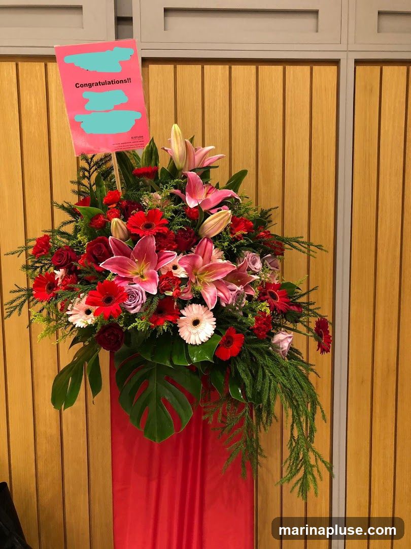 A large, vibrant floral arrangement featuring pink lilies, red roses, and gerbera daisies, accented with lush green foliage. A pink sign with "Congratulations!!" and obscured text is placed prominently among the flowers. The arrangement is displayed on a red draped stand against a backdrop of vertical wooden panels.