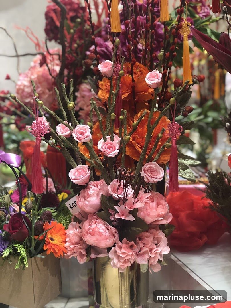 A close-up shot of a vibrant floral arrangement featuring numerous pink roses and orange flowers. The arrangement is adorned with decorative tassels in pink and orange, and some branches have small green buds. A price tag reading "$119" is visible on one of the pink flowers. The arrangement is displayed in a golden vase, with other floral displays and decorative items visible in the blurred background.