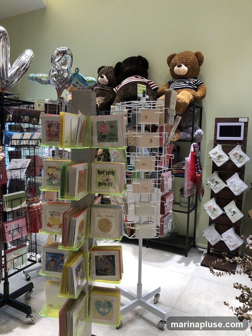 A display of greeting cards on a rotating stand, with various plush teddy bears and balloons in the background. There are also some small decorative items and tea bags on shelves to the right.