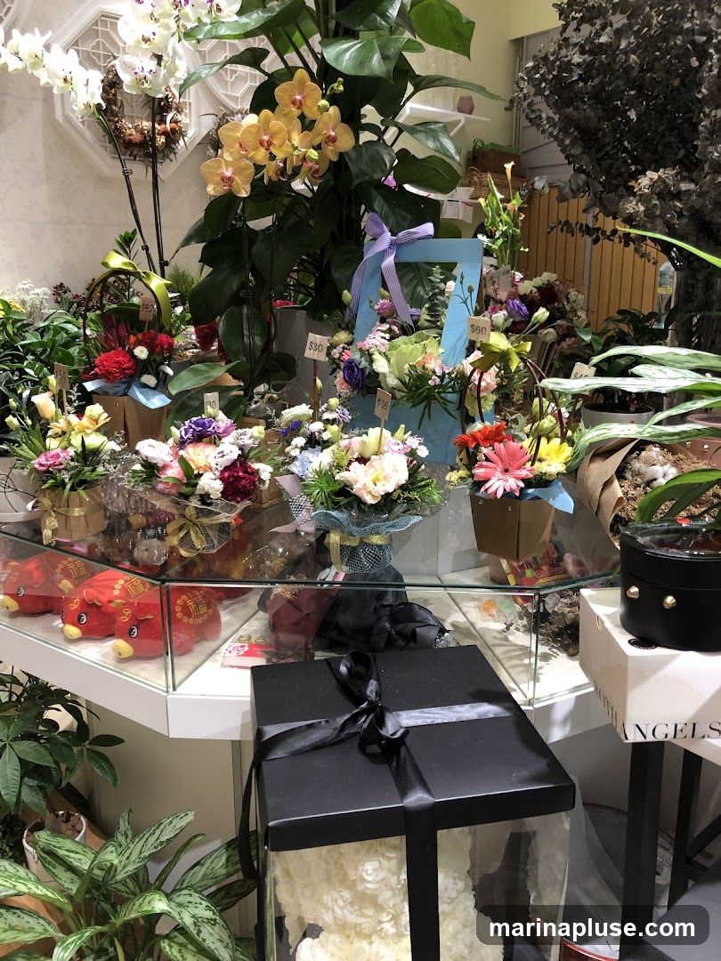 A display of various floral arrangements and plants inside a shop. In the foreground, a large black box with a ribbon contains a white floral bear. To the left, red pig-shaped plush toys are visible behind a glass counter. Various bouquets and potted plants are arranged on shelves and tables, with price tags indicating $15, $30, and $60. Orchids and other flowering plants are prominently featured.