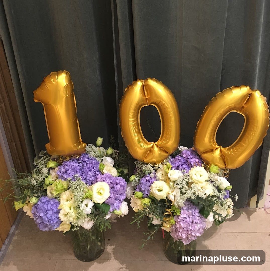 Two large floral arrangements are displayed in glass vases. Each arrangement features a mix of purple hydrangeas, white and cream roses, and various green foliage. Gold Mylar balloons in the shape of the numbers