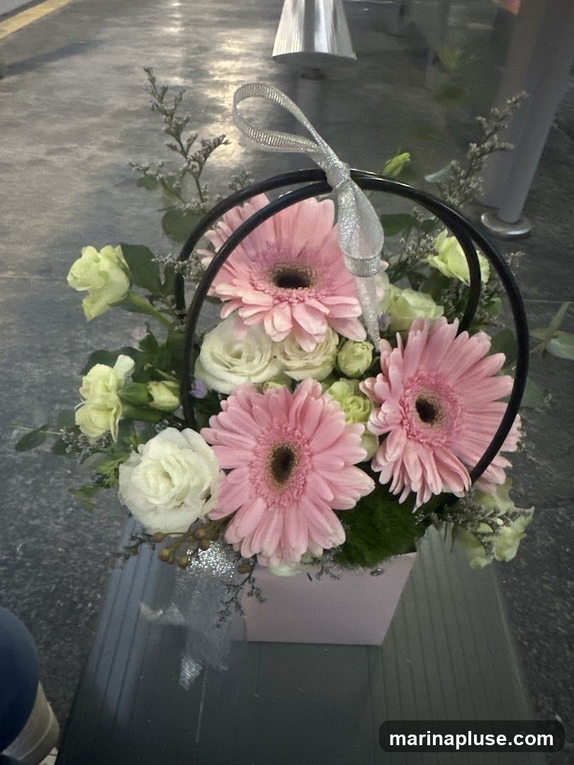 A pink square box holds a floral arrangement featuring pink gerbera daisies, white roses, and light green flowers. The arrangement is adorned with a silver ribbon tied in a bow and a black metal hoop. The background appears to be an outdoor setting with a textured gray surface.