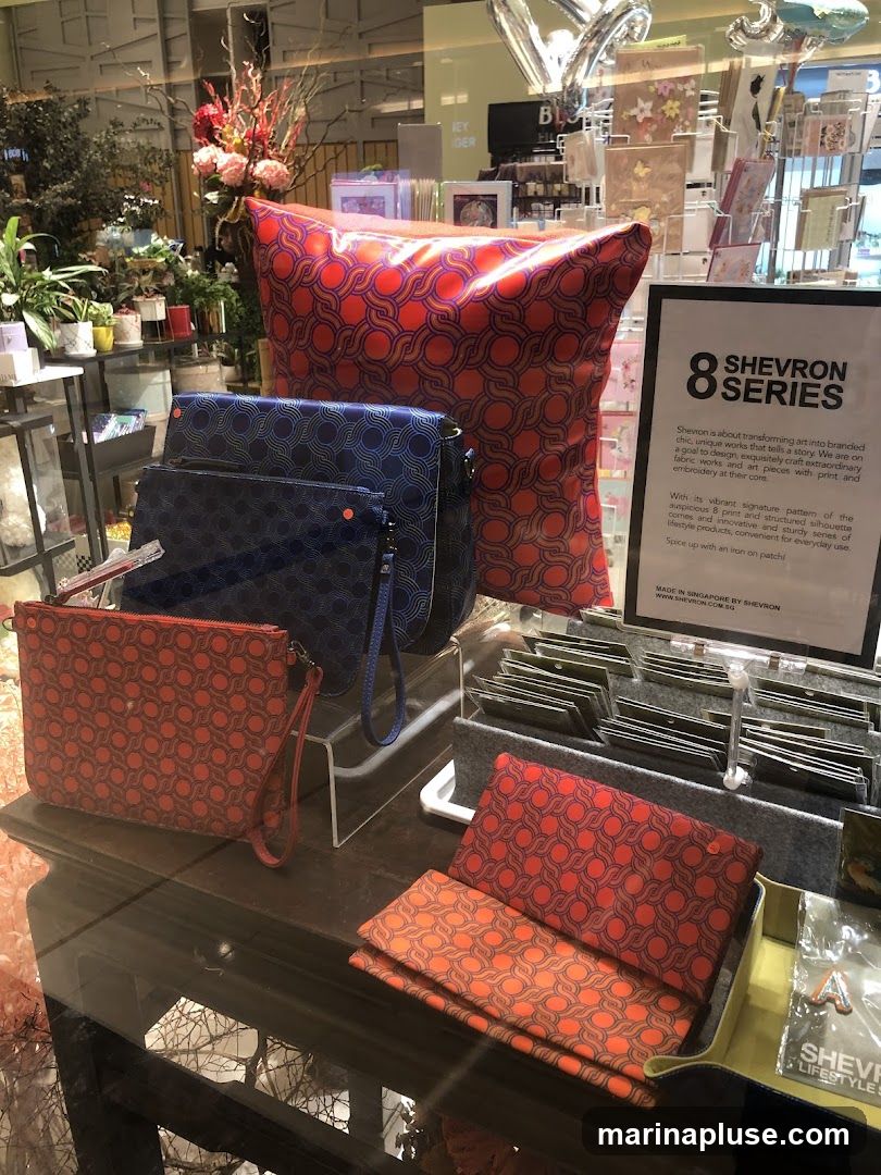A display of various lifestyle products featuring the "8 Chevron Series" pattern. Items include a large cushion, several pouches and clutches in blue and red, and folded fabric items. A sign describes the "Chevron Series" as transforming art into branded chic works, highlighting the vibrant signature pattern and structured silhouettes. The products are made in Singapore by Shevron.