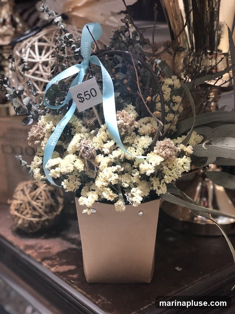 A close-up of a dried flower arrangement in a light brown paper bag. The arrangement features small white and beige dried flowers, along with eucalyptus leaves and twigs. A light blue ribbon with the word "BLOSSOM" printed on it is tied around the flowers, and a price tag reading "$50" is attached. In the background, a decorative woven sphere and a metallic vase are visible.