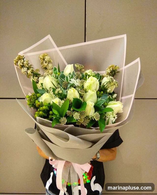 A person holds a large bouquet of white roses and other white and green flowers, wrapped in grey and translucent paper with a pink ribbon.