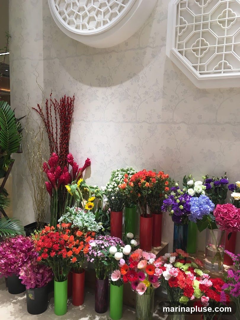 A vibrant display of various flowers in vases and buckets, arranged against a patterned wallpaper. The flowers include red ginger, sunflowers, roses, hydrangeas, and gerbera daisies in a multitude of colors.