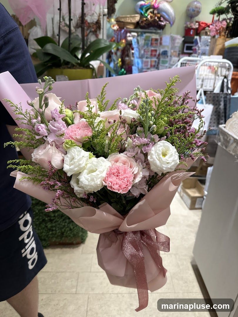 A person is holding a bouquet of pink and white flowers wrapped in light pink paper and tied with a rose gold ribbon. The background appears to be a flower shop with shelves of plants, balloons, and other floral arrangements.