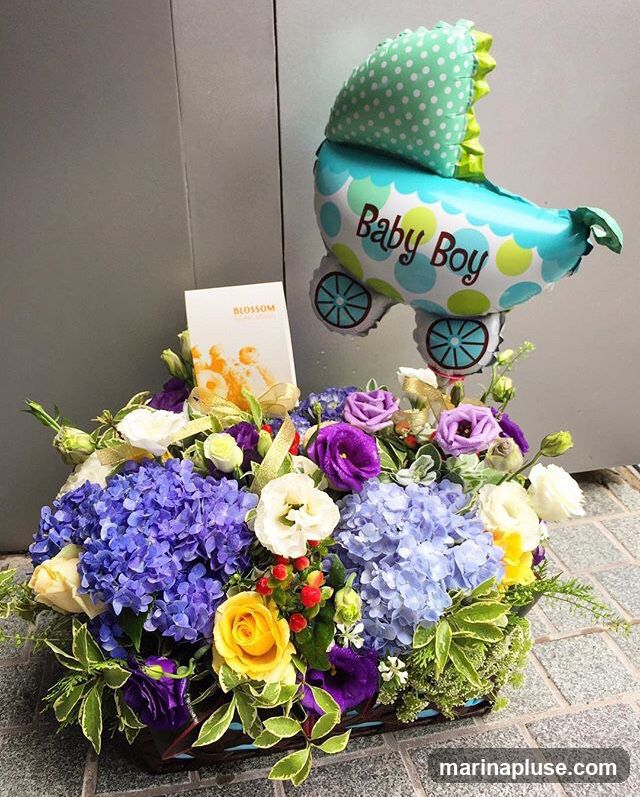 A basket arrangement of flowers in shades of purple, blue, yellow, and white, with a "Baby Boy" balloon shaped like a stroller. A card with the word "BLOSSOM" is also visible.
