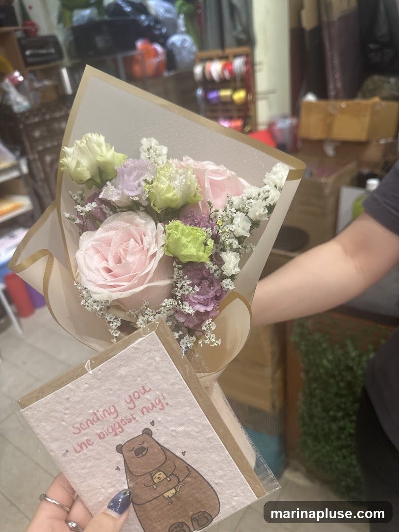 A person is holding a bouquet of pink and green roses with small white and purple flowers, wrapped in translucent paper. Below the bouquet, they are holding a greeting card with a drawing of a bear hugging a smaller bear and the text "Sending you the biggest hug!". The background appears to be a shop with shelves stocked with various items, including rolls of ribbon and boxes.