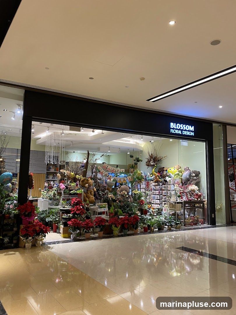 A well-lit floral shop with a wide display of plants, flowers, teddy bears, and other gift items. The shop's name, "BLOSSOM FLORAL DESIGN," is visible on a sign above the entrance.