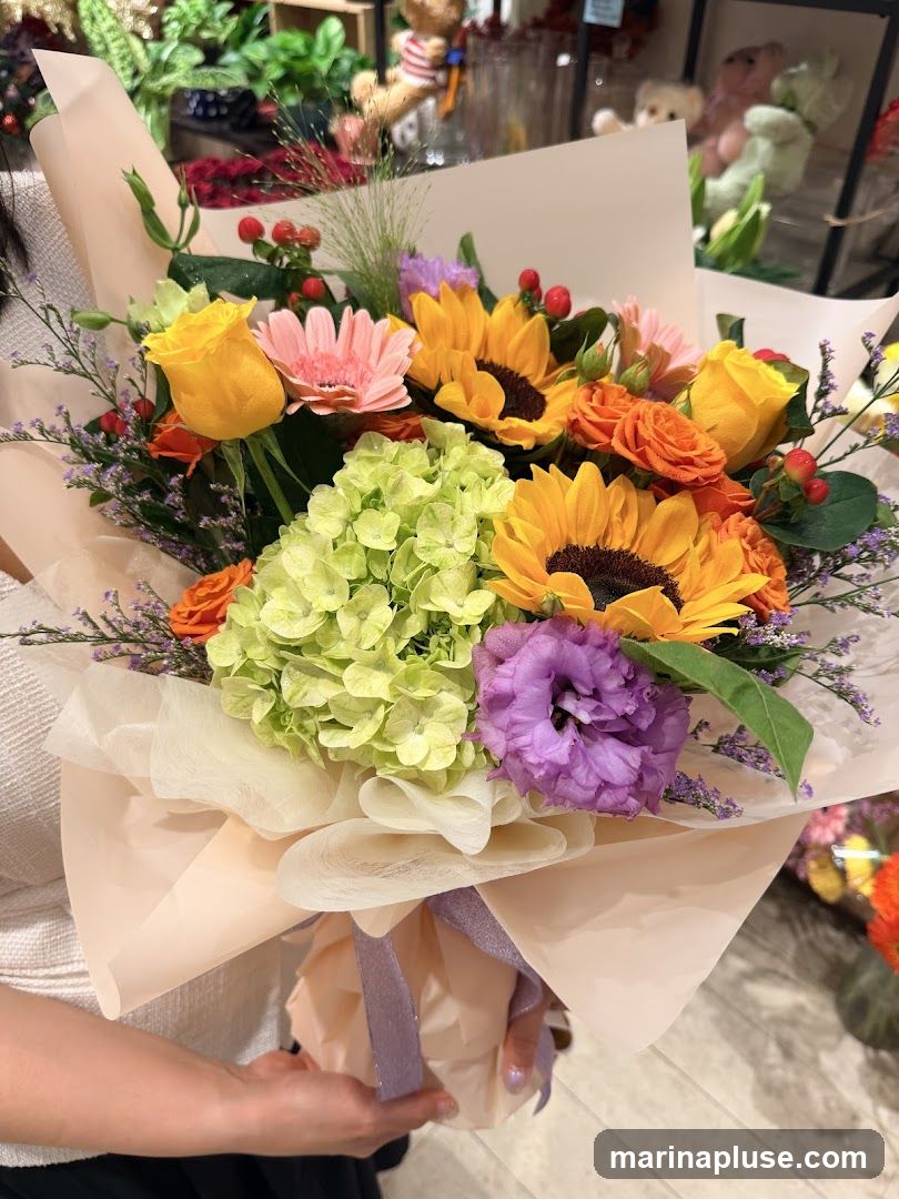 A close-up shot of a person holding a large, colorful bouquet of flowers. The bouquet features sunflowers, orange roses, yellow roses, a pink gerbera daisy, light green hydrangeas, and purple lisianthus, accented with small red berries and delicate purple filler flowers. The arrangement is wrapped in pale peach and cream paper, tied with a shimmering lavender ribbon. In the blurred background, shelves with decorative items like teddy bears and plants are visible, suggesting a retail environment such as a flower shop.