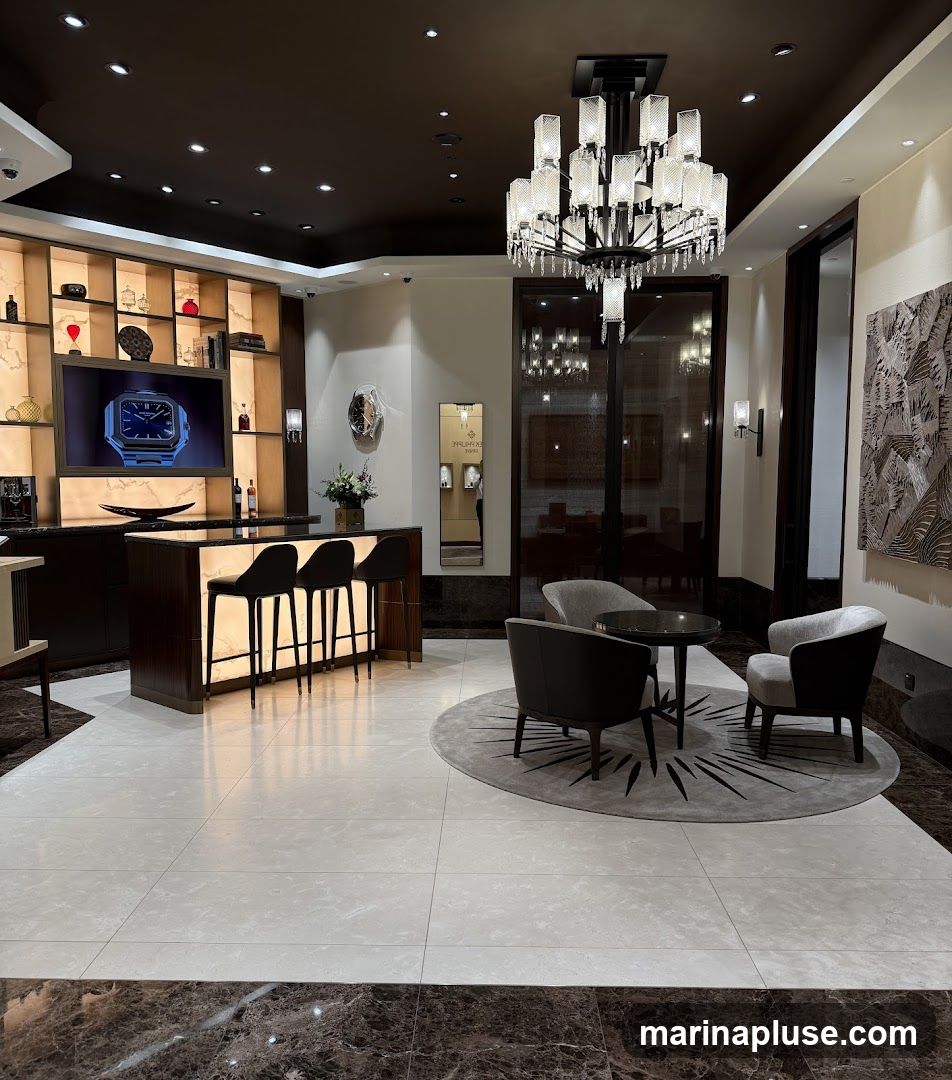 The interior of a high-end watch store features a modern bar area with illuminated stools, a large screen displaying a watch, and shelves stocked with various items. A seating area with plush chairs and a coffee table is arranged on a circular rug. A large, ornate chandelier hangs from the dark ceiling, and artwork adorns the walls.