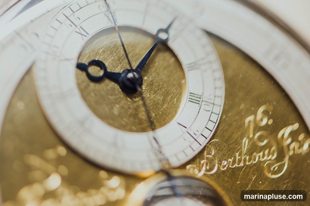 A detailed, close-up shot of a vintage pocket watch face. The watch features a white dial with Roman numerals and intricate gold-toned metalwork. The hands of the watch are visible, indicating a time around 10:50. Engraved on the gold surface are the words 'Berthous Frie' and the number '76'. The image has a shallow depth of field, with the foreground and background blurred, drawing focus to the central details of the watch.