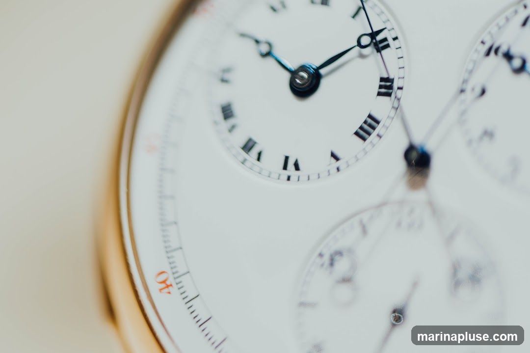 A detailed, close-up shot of a white watch face with multiple subdials. The main dial features Roman numerals, with hands pointing to approximately 10:10. Smaller subdials are visible, one with Arabic numerals and another with what appears to be a minute or second counter. The watch has a gold-toned bezel and a light-colored background, suggesting a studio product shot.