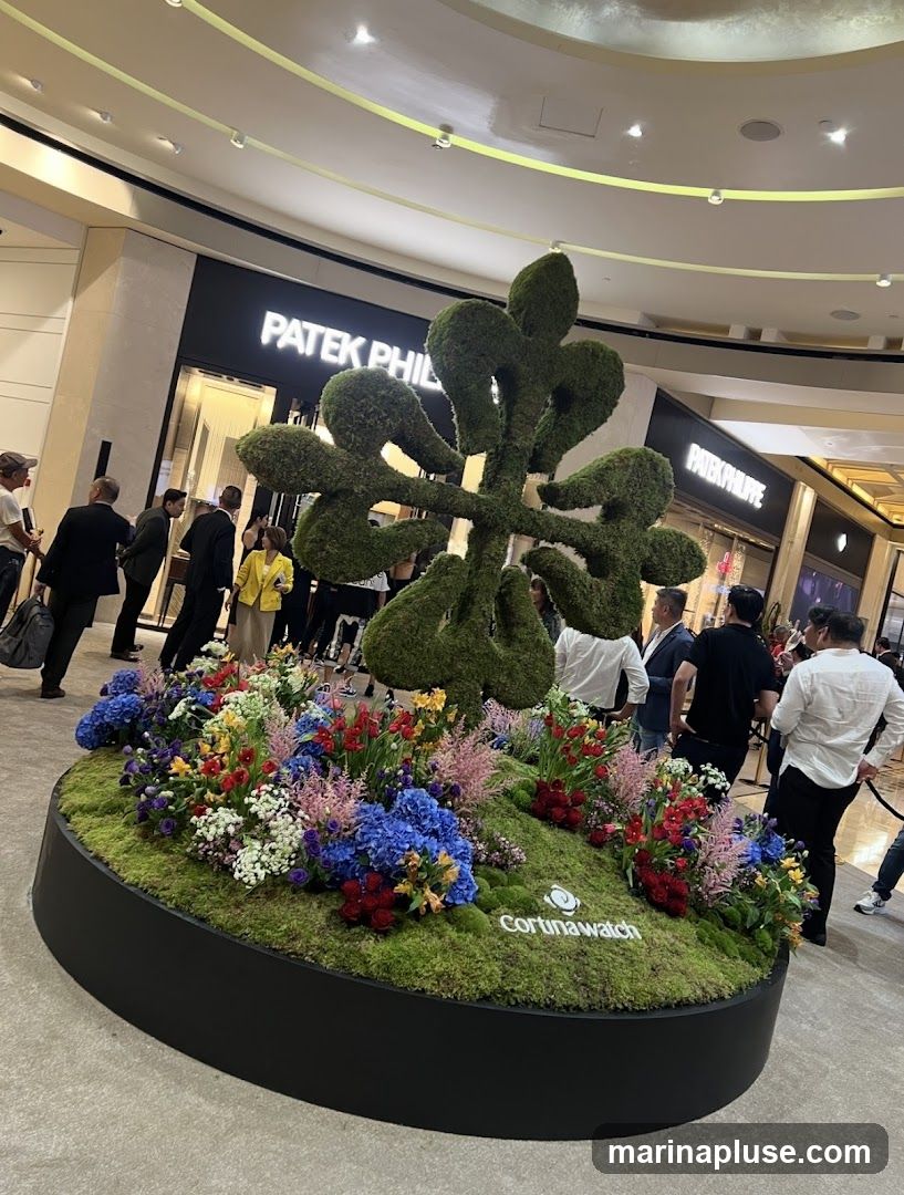 A large, sculpted green topiary resembling Chinese characters stands in the center of a circular display of colorful flowers and moss. The display has a black base with the "Cortinawatch" logo visible. In the background, the storefronts of "PATEK PHILIPPE" are visible, with people walking by.