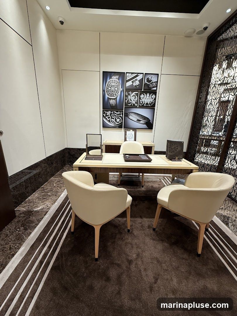 A well-appointed seating area within a luxury watch boutique, featuring a large desk with a framed display of intricate watch designs and close-ups of watch components. Three plush, cream-colored armchairs are arranged around the desk, inviting customers to sit and discuss purchases. The flooring is a combination of polished marble and a plush brown carpet with a striped accent. A decorative metal screen is visible on the right side of the image, adding to the sophisticated ambiance.