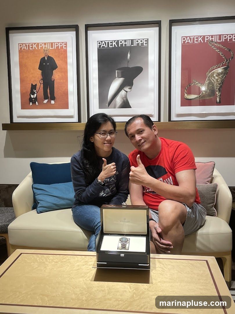 A man and a woman are sitting on a couch, both giving a thumbs up. In front of them, on a table, is an open watch box containing a Patek Philippe watch. Behind them, three framed Patek Philippe advertisements are displayed on the wall.
