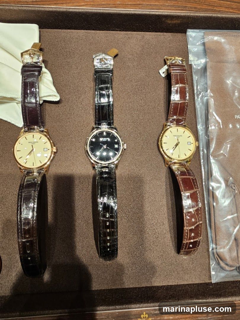 Three luxury watches with leather straps are displayed on a brown textured surface. The watches have gold cases and different colored dials. One watch has a cream-colored dial with Roman numerals, another has a black dial, and the third has a cream-colored dial with stick markers. A white cloth is visible to the left of the watches, and a brown leather pouch is to the right.