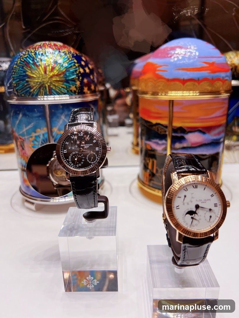 Two luxury watches with black leather straps are displayed on clear acrylic stands. The watch on the left has a black dial with Roman numerals and sub-dials. The watch on the right has a white dial with Roman numerals and a moon phase complication. Behind the watches are decorative cylindrical objects with colorful, artistic designs, possibly music boxes or display elements. The background is blurred, suggesting an indoor display setting, likely a jewelry or watch store.