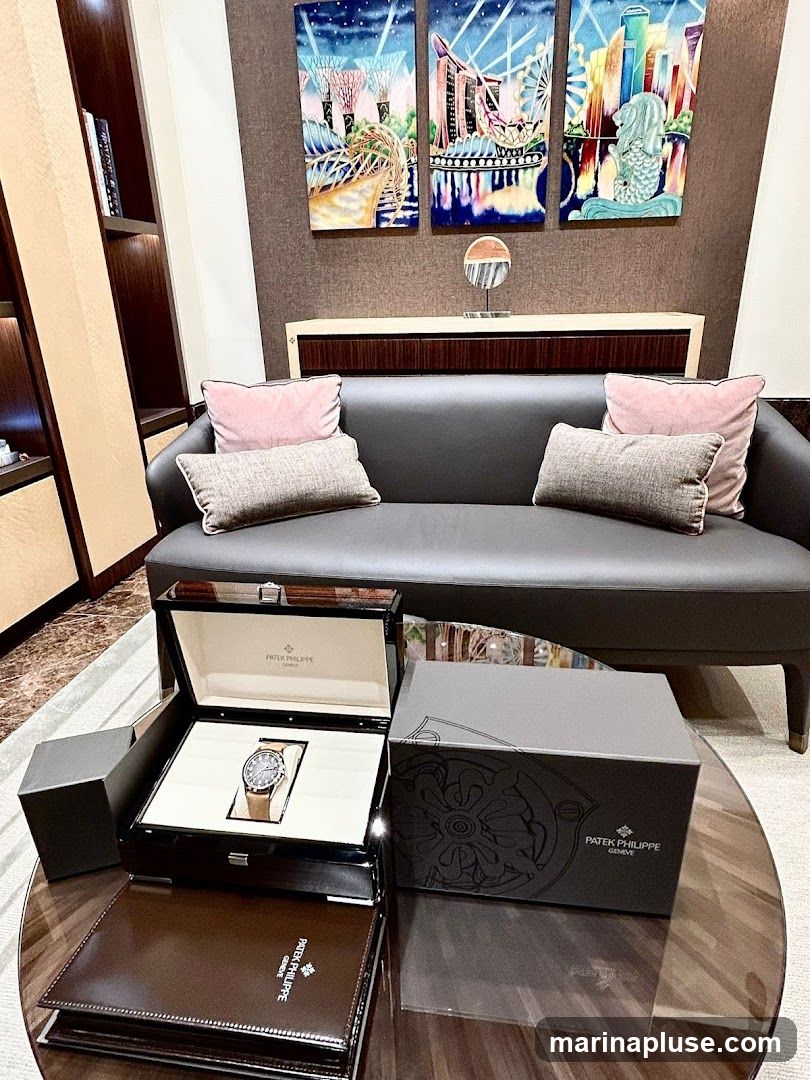 A Patek Philippe watch is displayed in an open wooden box on a glass table. Next to it is a closed Patek Philippe box and a leather-bound folder. The background features a dark grey sofa with decorative pillows, and a triptych artwork depicting Singapore landmarks above a console table. Built-in shelving is visible on the left.