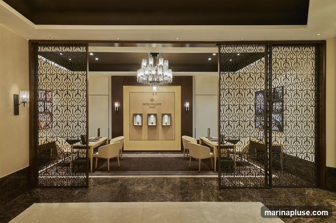 A luxurious showroom interior with ornate screens, plush seating, and display cases. The Patek Philippe Geneva logo is prominently displayed above three illuminated display niches. A large chandelier hangs from the ceiling, and elegant wall sconces provide additional lighting. The flooring is a rich marble.