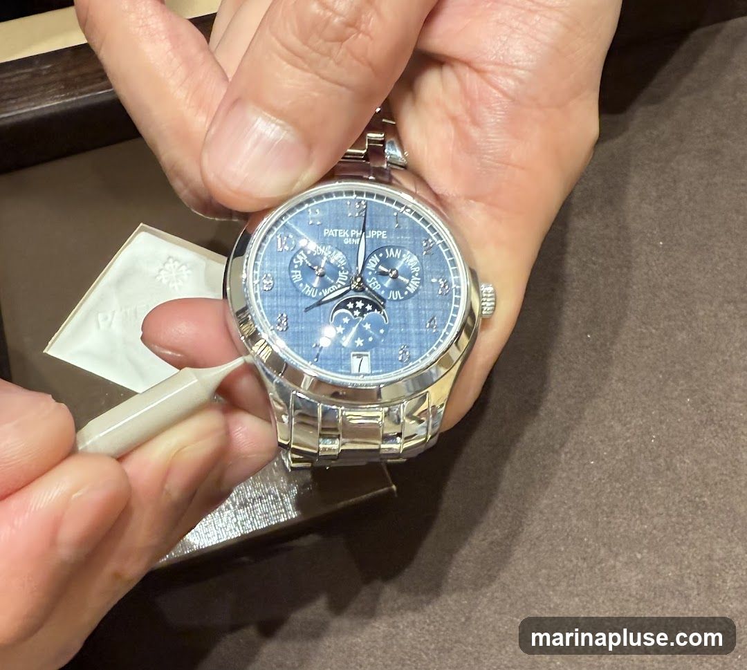 A close-up shot of a person's hands adjusting a Patek Philippe watch with a blue dial and moon phase complication. The watch has a stainless steel bracelet. A white cloth with the Patek Philippe logo is visible to the left.