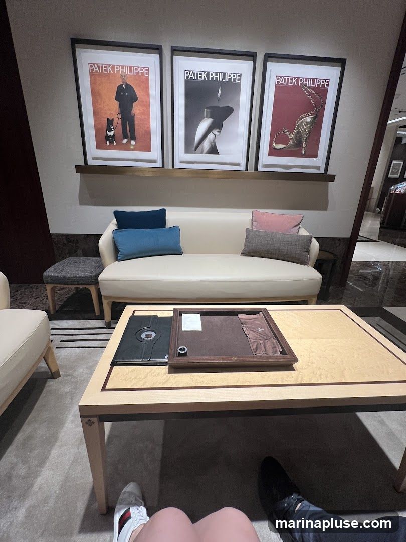 A seating area with a sofa and a coffee table, featuring framed Patek Philippe advertisements on the wall. The coffee table has a display tray with watch accessories.