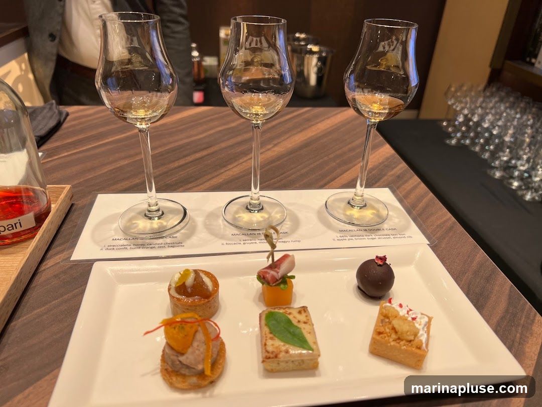 Three tasting glasses filled with amber liquid are arranged on a white card with text. In front of the glasses, a white rectangular plate holds six small appetizers. The appetizers include a tart with fruit, a bite-sized pastry with meat and orange zest, a square pastry with a green leaf, a chocolate truffle topped with a rose petal, and a small tart with white cream and red sprinkles. A bottle with red liquid is visible to the left. In the background, blurred glasses and a bar area are visible.