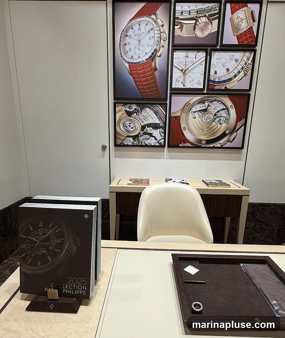 A display area with framed images of watches and watch mechanisms on the wall. In the foreground, there are stacks of brochures titled "2025 COLLECTION PATEK PHILIPPE" and a display tray with watch accessories. A cream-colored chair is positioned behind a desk.