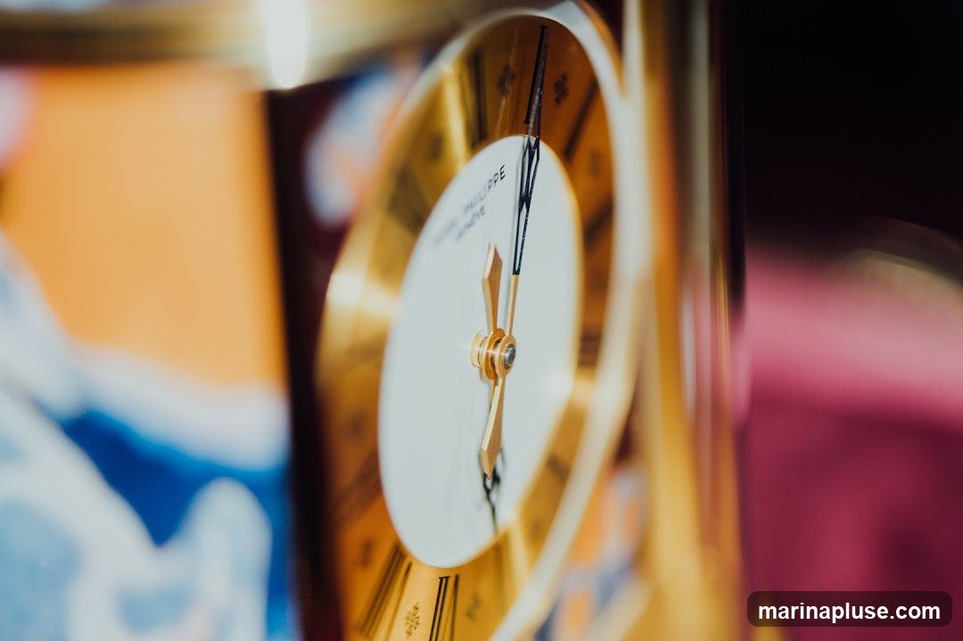A close-up, shallow depth-of-field shot of a golden clock face with Roman numerals. The hands of the clock are visible, pointing to approximately 10:08. The brand name 'Patek Philippe Geneve' is partially visible on the dial. The background is blurred, showing hints of orange, blue, and dark red.