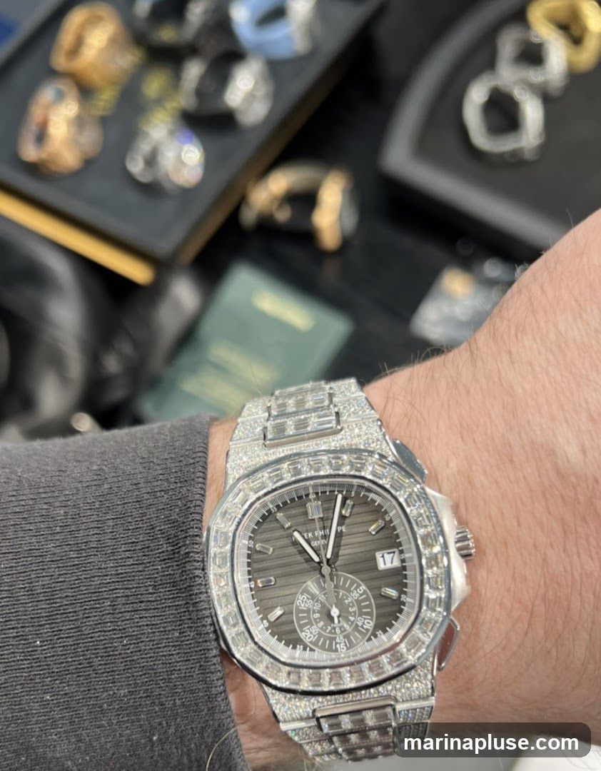 A close-up shot of a person's wrist wearing a diamond-encrusted watch. The watch has a dark grey dial with silver hands and hour markers. A date window is visible at the 3 o'clock position, and a chronograph subdial is present at the 6 o'clock position. The watch band and bezel are also covered in diamonds. In the blurred background, various pieces of jewelry, including rings and bracelets, are displayed on black trays.
