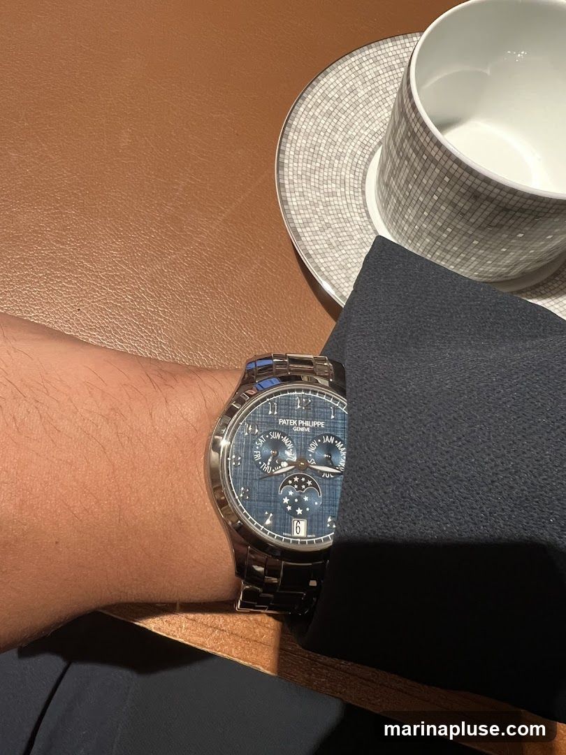 A close-up shot of a person's wrist wearing a Patek Philippe watch with a blue textured dial. The watch features multiple subdials and a date window. Next to the watch is a white coffee cup and saucer with a mosaic-like pattern. The setting appears to be a table with a brown leather surface.