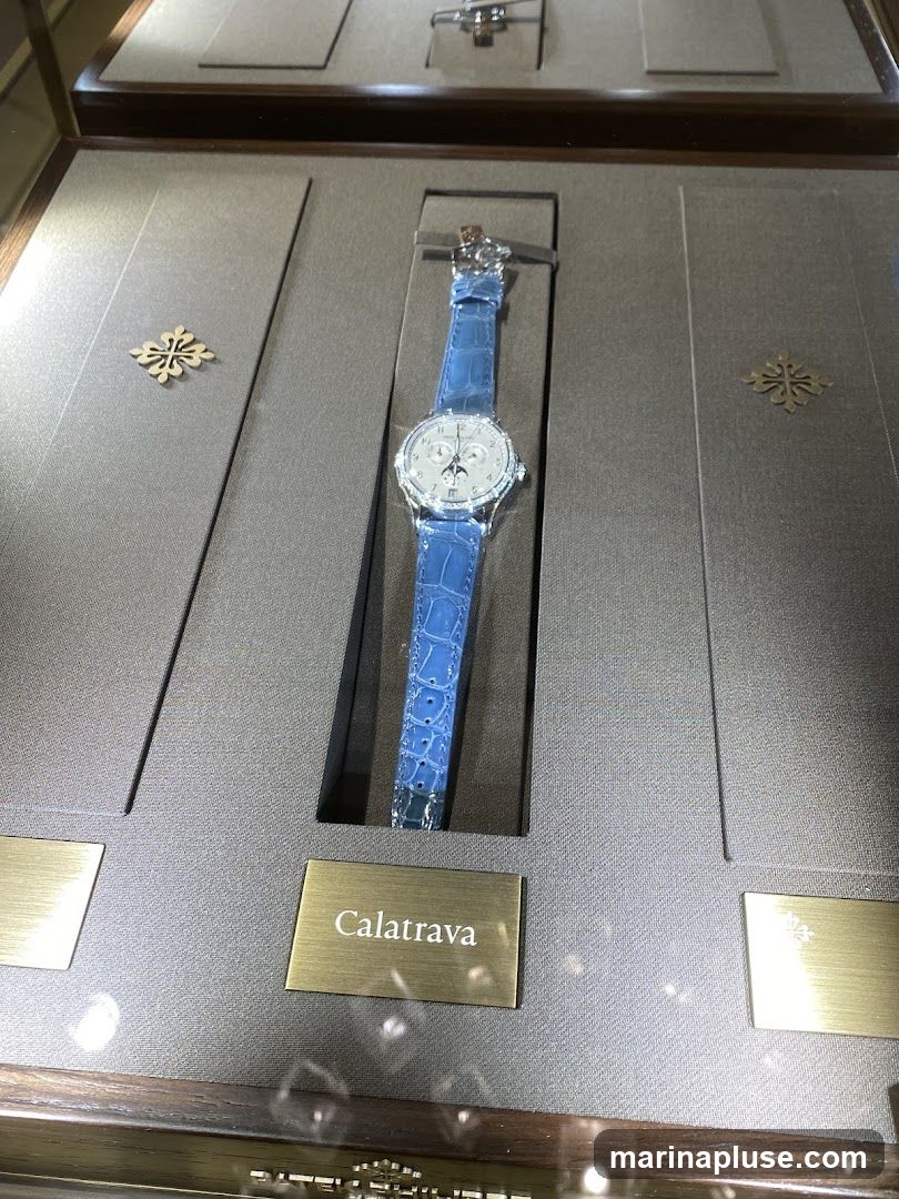 A luxury watch with a blue leather strap is displayed in a glass case. The watch face is silver with intricate details and a moon phase complication. A plaque below the watch reads "Calatrava". The display case has a textured gray background with decorative golden emblems.