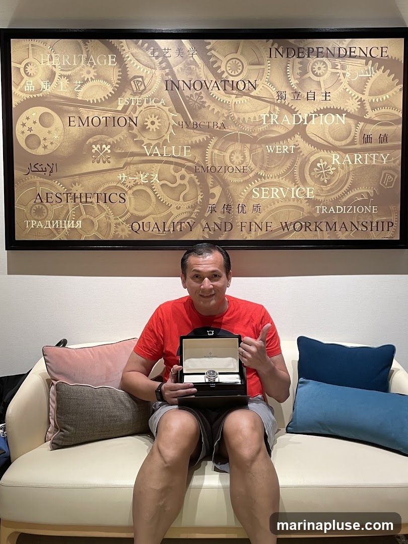 A man in a red t-shirt and grey shorts sits on a cream-colored couch, holding an open watch box with a watch inside. He is giving a thumbs-up gesture. Behind him is a large framed artwork featuring gears and various words in different languages, including "HERITAGE," "INNOVATION," "EMOTION," "VALUE," "AESTHETICS," "QUALITY AND FINE WORKMANSHIP," "INDEPENDENCE," "TRADITION," "WERTH," "RARITY," and "SERVICE."