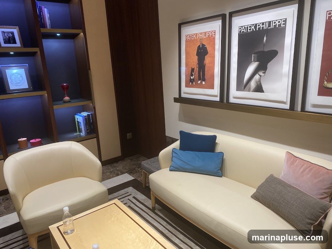 A comfortable and elegant lounge area with a cream-colored sofa and armchair. The sofa is adorned with several decorative pillows in shades of blue, pink, and grey. A low, rectangular coffee table with a golden frame sits in front of the sofa. Behind the seating area, a modern bookshelf is illuminated with soft lighting, displaying books and decorative items. The wall above the sofa features framed Patek Philippe advertisements, showcasing the brand's sophisticated aesthetic.