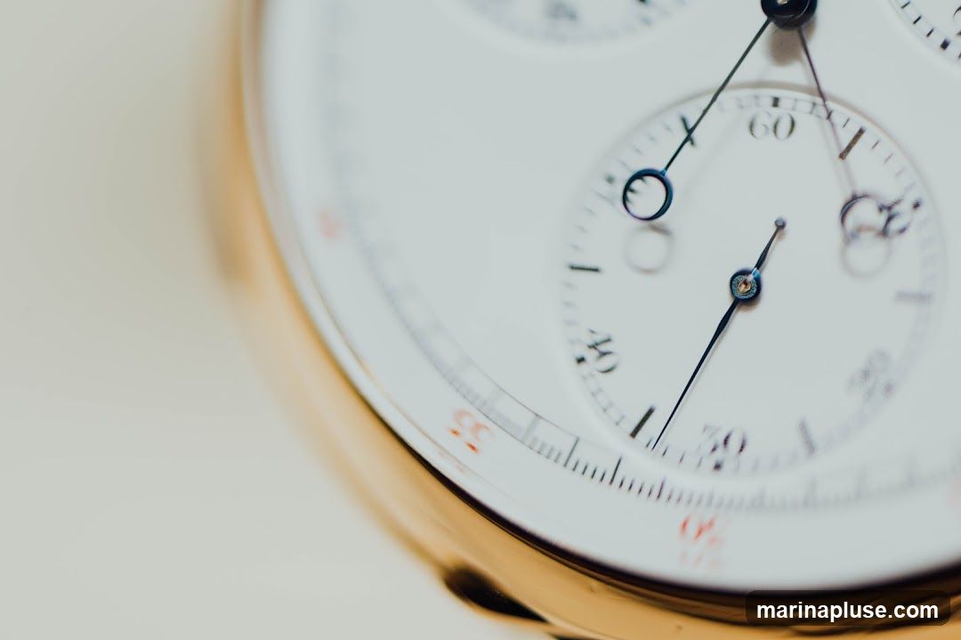 A detailed, shallow depth-of-field shot focuses on the white face of a gold-cased chronograph watch. The image highlights the intricate sub-dials and hands, with markings for minutes and seconds visible. The primary hands are dark, contrasting with the white face, and the gold bezel of the watch is softly blurred in the foreground and background.