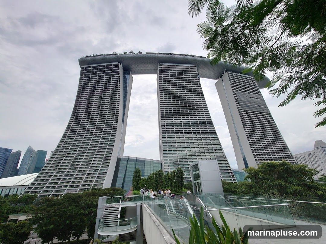 A wide-angle shot of the Marina Bay Sands hotel in Singapore, with its distinctive three towers connected by a sky-high platform. The hotel is viewed from a slightly elevated perspective, with lush greenery and a walkway in the foreground. The sky is overcast, and other buildings are visible in the distance.