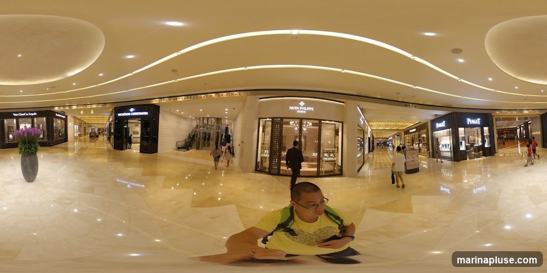 A wide-angle view of a brightly lit, upscale shopping mall interior. Several luxury watch and jewelry stores are visible, including Van Cleef & Arpels, Vacheron Constantin, Patek Philippe, and Piaget. Shoppers are seen walking through the polished marble-floored corridors. A man in a yellow shirt is in the foreground, taking a selfie.