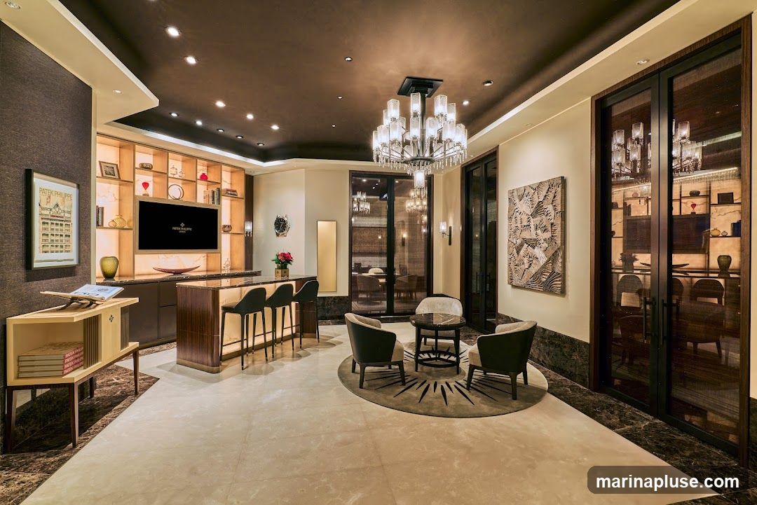 A high-end watch boutique interior featuring a modern bar area with stools, a seating lounge with armchairs and a coffee table, and display shelves. A large television screen displays the Patek Philippe logo. Artwork adorns the walls, and a prominent chandelier hangs from the ceiling. The overall ambiance is sophisticated and elegant.