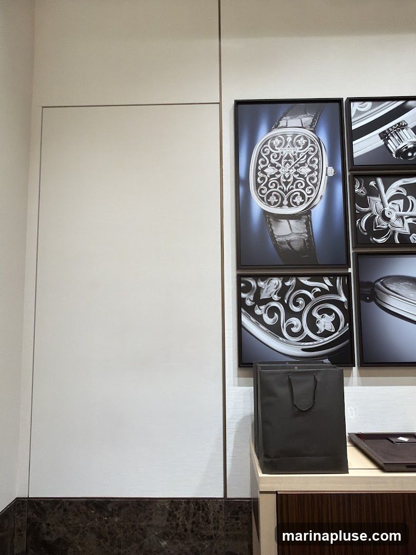 A display featuring framed images of a luxury watch and intricate watch components. A dark shopping bag sits on a counter below the images, with a display tray to its right. The wall behind the display is plain and light-colored.