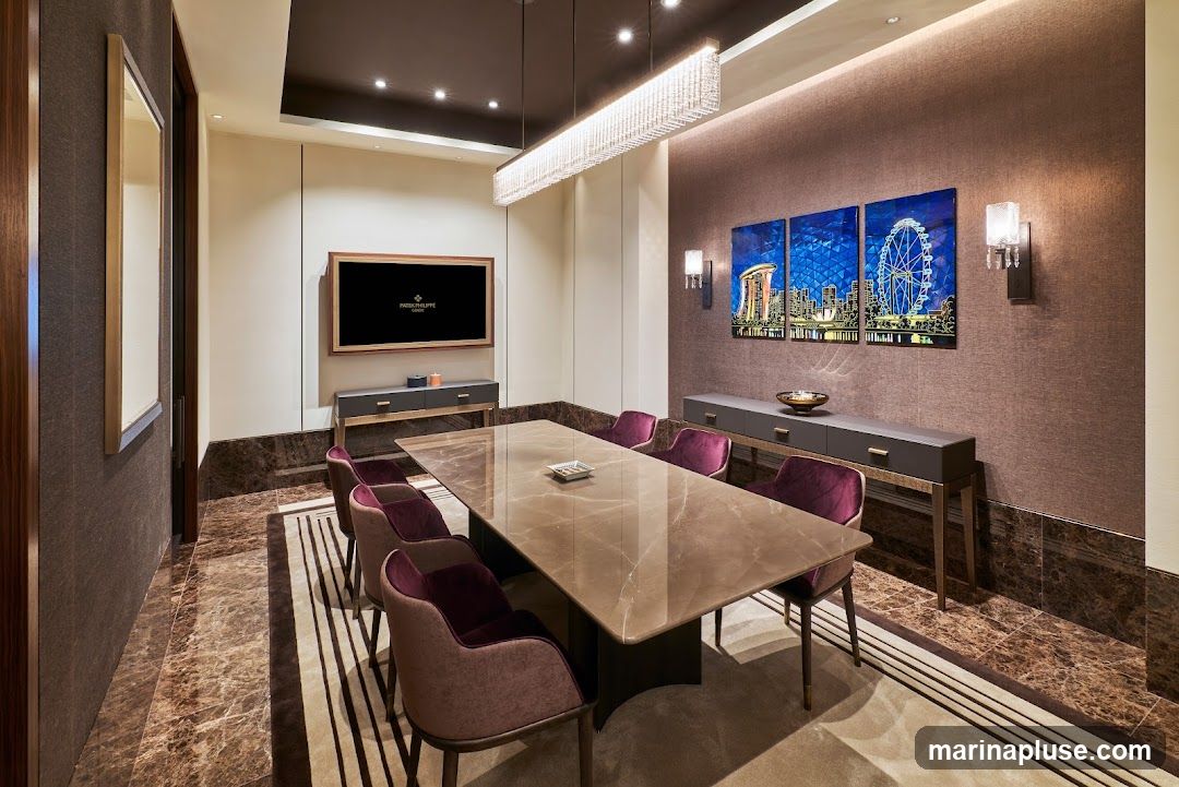 A sophisticated meeting room features a long, polished stone table surrounded by plush purple chairs. A large television screen on the wall displays the Patek Philippe logo. Three vibrant artworks depicting a cityscape with a Ferris wheel adorn the textured brown wall. The room is illuminated by a modern chandelier and wall sconces, with marble flooring and a patterned rug adding to the luxurious ambiance.